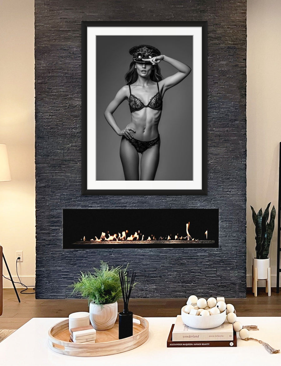 Framed fine art photo hanging above a fireplace in a stylish casual livingroom showing a black and white image of a gorgeous woman wearing a lace bra and panties with a steampunk style captain's hat and round goggles on her head with one hand on hip and the other saluting the camera.