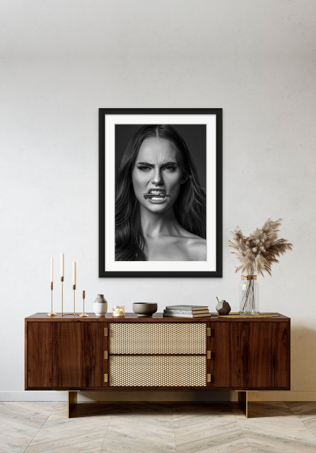 Framed artwork hanging above a brown walnut credenza, the art is a black and white close up portrait of a beautiful woman holding a bullet between her teeth with her brow furrowed.