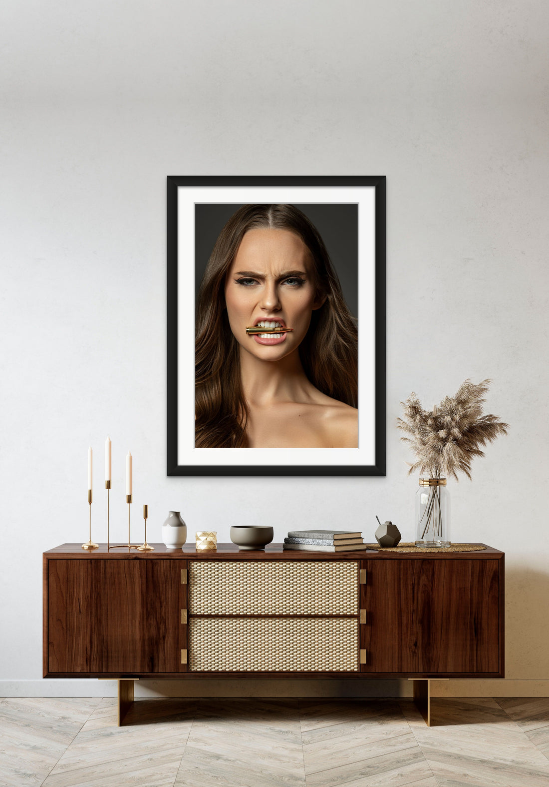 Framed artwork hanging above a brown walnut credenza, the art is a close up portrait of a beautiful woman holding a bullet between her teeth with her brow furrowed.