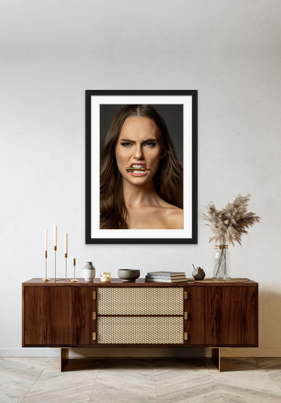 Framed artwork hanging above a brown walnut credenza, the art is a close up portrait of a beautiful woman holding a bullet between her teeth with her brow furrowed.