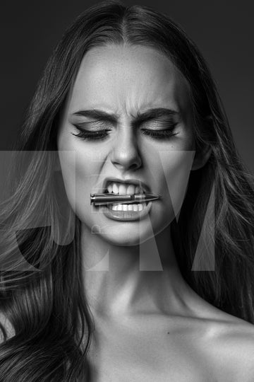 Close up black and white portrait of a beautiful woman holding a bullet between her teeth with her brow furrowed.