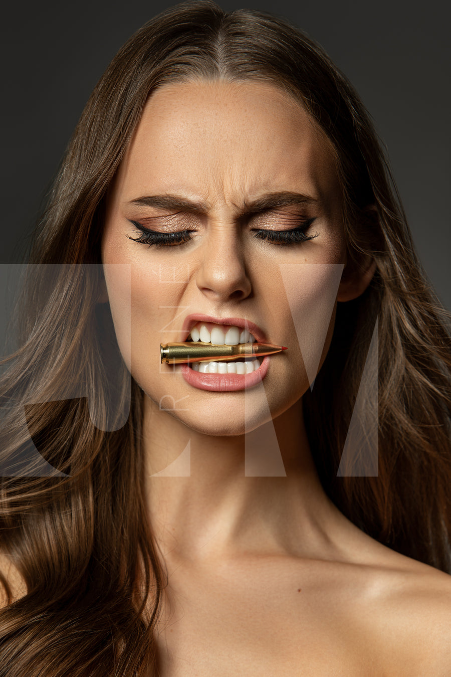 Close up portrait of a beautiful woman holding a bullet between her teeth with her brow furrowed.