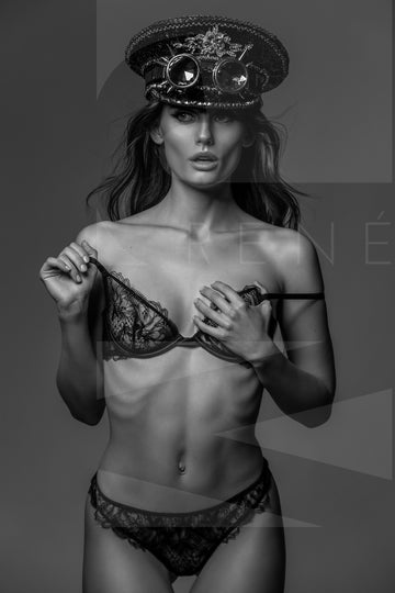 Black and white photo of a gorgeous woman wearing a lace bra and panties with a steampunk style captain's hat and round goggles on her head. 