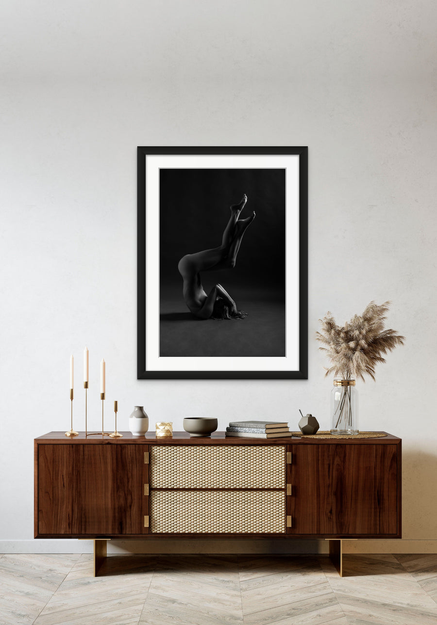 Framed artwork hanging above a brown walnut credenza, the art is a black and white photo showing a sideview of a naked woman in a shoulder stand with arms bent covering her face and knees bent with legs crossed.