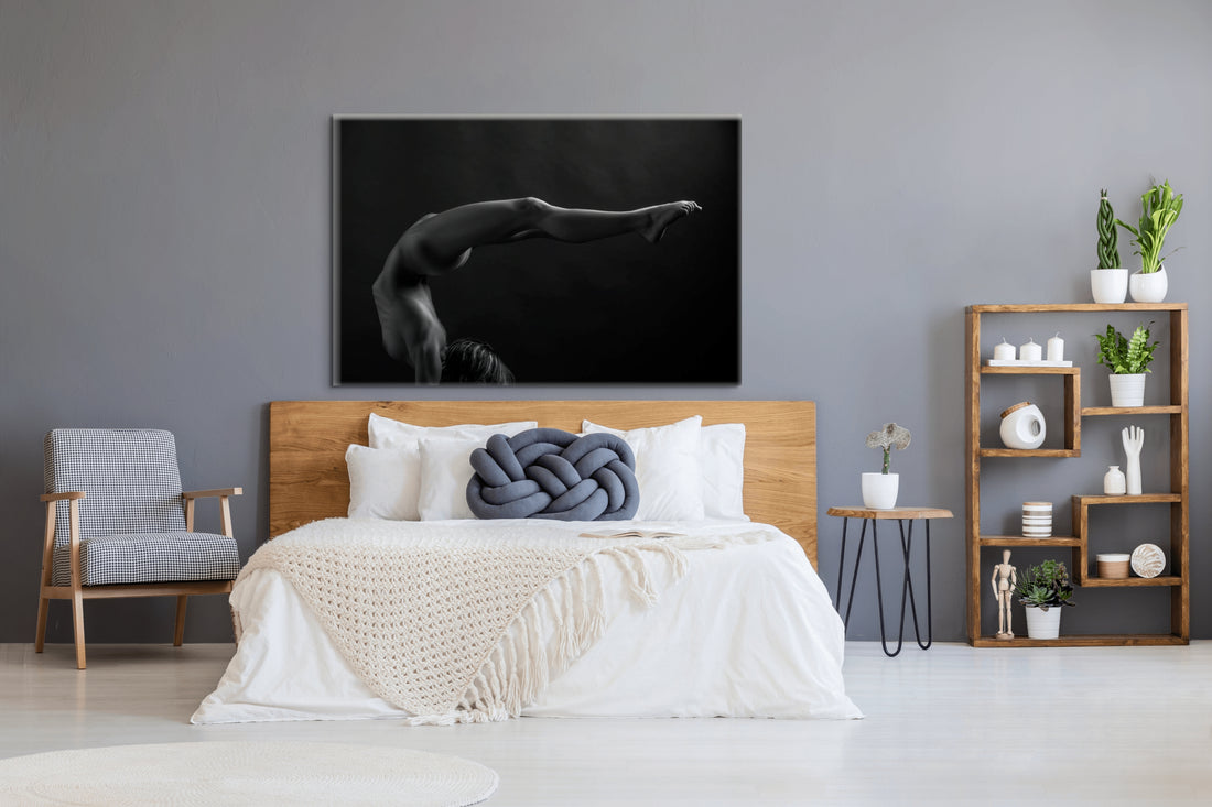 Canvas artwork hanging above a light coloured wood headboard in a bedroom with natural and blue tones, the art is a black and white photograph of a sideview of a nude woman in a handstand pose with back arched in a sweeping curve with legs stretching out behind her.