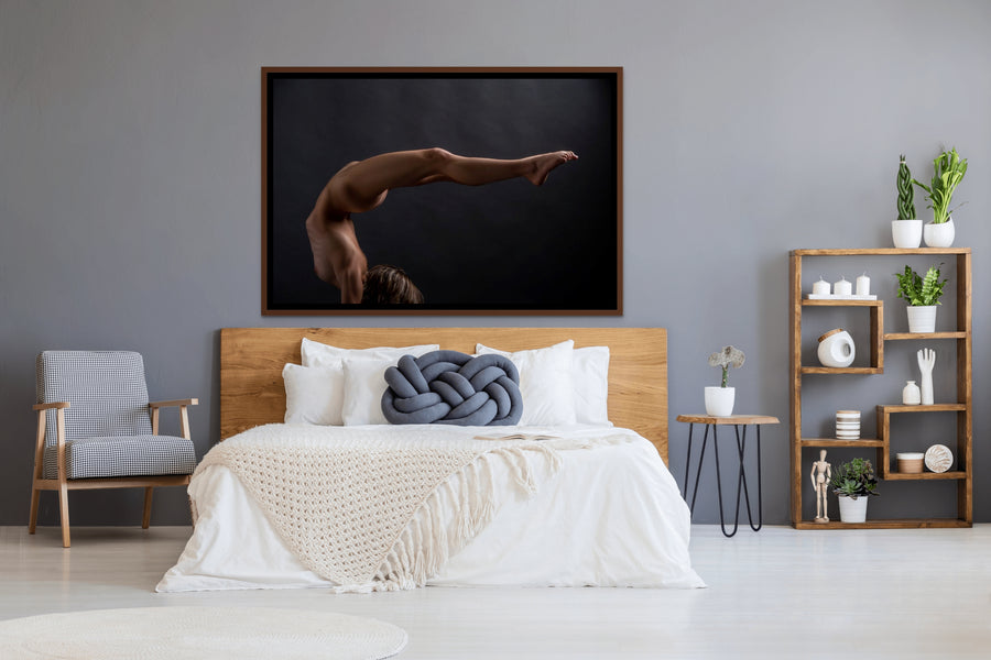 Framed canvas artwork hanging above a light coloured wood headboard in a bedroom with natural and blue tones, the art is a photograph of a sideview of a nude woman in a handstand pose with back arched in a sweeping curve with legs stretching out behind her.