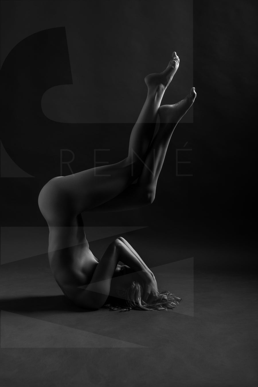 Black and white photo showing a sideview of a naked woman in a shoulder stand with arms bent covering her face and knees bent with legs crossed.