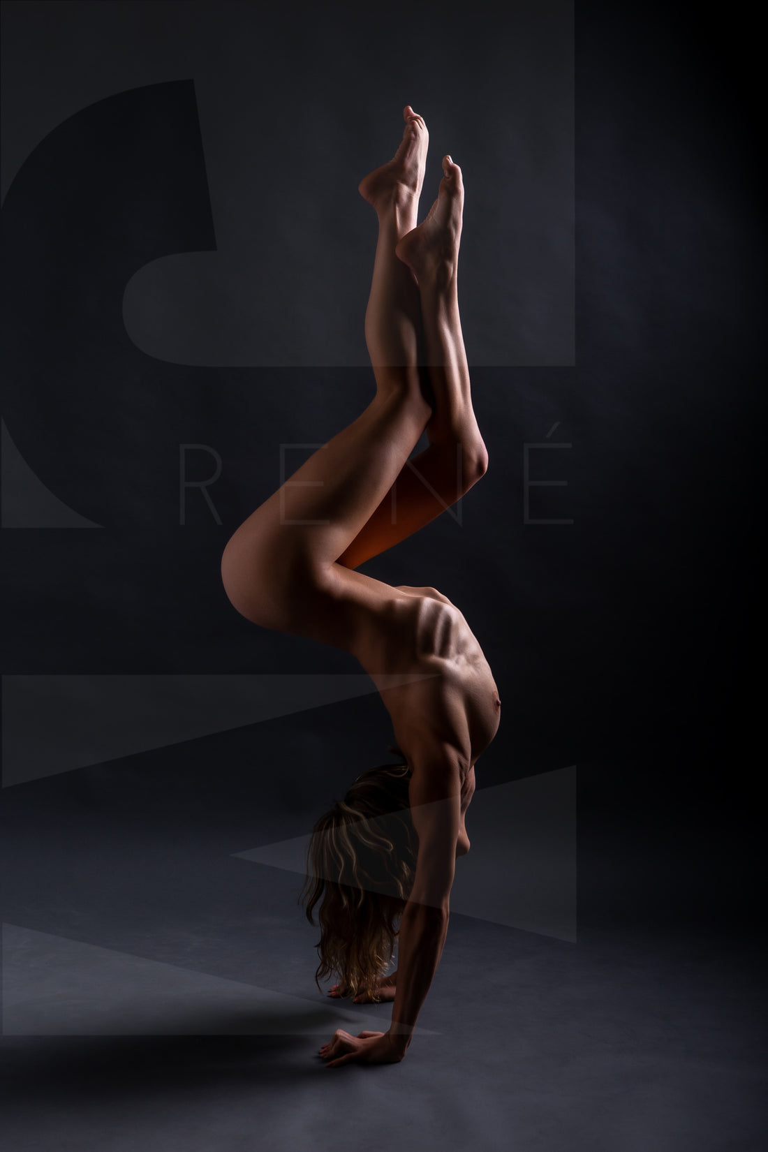 A side body view of a nude woman in a handstand pose with arched back and crossed bent legs.