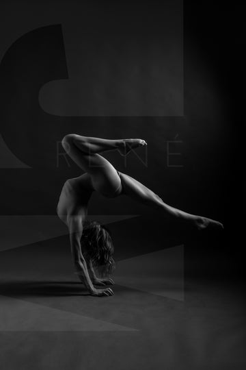 Black and white photo showing a sideview of a nude woman in a handstand pose, balance with one leg pointed straight back behind the woman and the other knee bent directly above the woman.