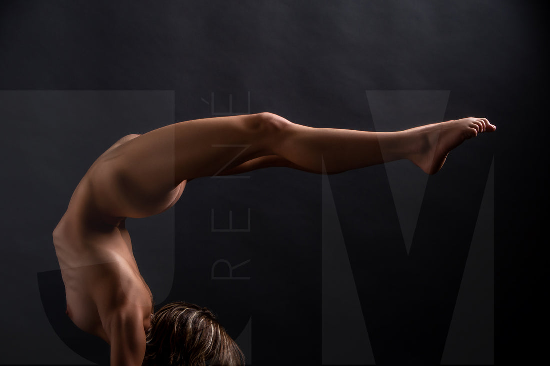 A sideview of a nude woman in a handstand pose with back arched in a sweeping curve with legs stretching out behind her.