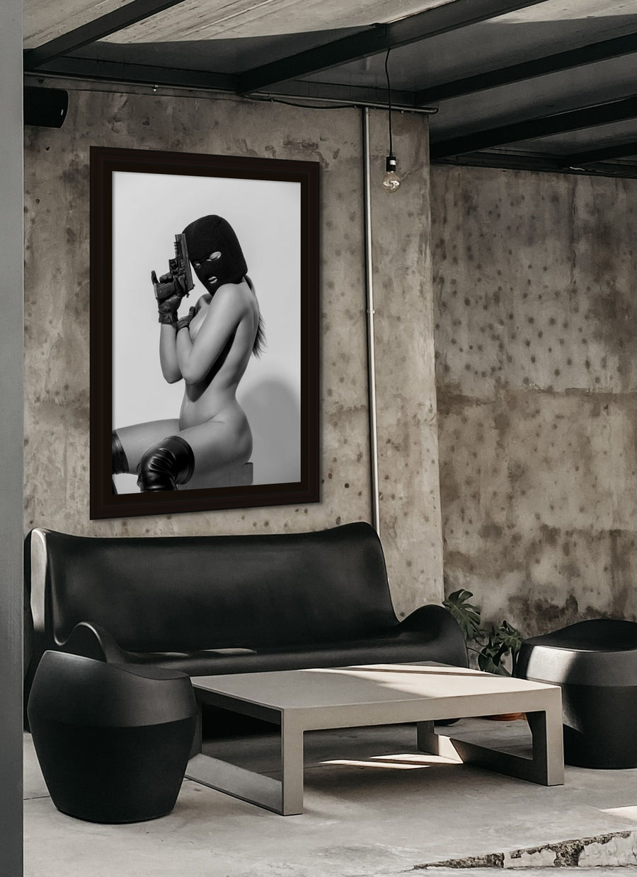 Framed canvas artwork hanging above a black leather couch in an industrial style living room, the artwork is a photo of a seated nude woman, the woman is wearing black thigh high boots, a black balaclava, black leather gloves and is holding a handgun.